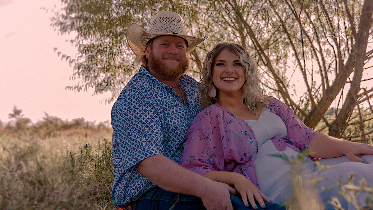 Smiling couple sitting together outdoors in a grassy field, with the man wearing a cowboy hat and patterned shirt, and the woman in a light dress and floral shawl. Sunlit trees and warm, natural light in the background.