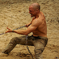Shirtless muscular man pulling a heavy chain with effort on sandy ground, outdoors, showing strength and determination in action.