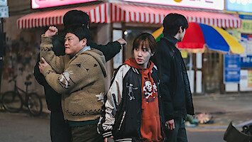 Four people stand on a city street at night near shops with striped awnings; one person looks concerned, two face away, and the street is dimly lit with visible store signs and urban elements.