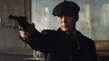 A serious man wearing a flat cap and suit points a handgun in a dimly lit room with vintage decor, suggesting a tense confrontation or dramatic moment.