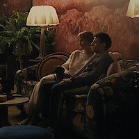 A couple sits closely together on a floral sofa with a small dog in a cozy, dimly lit living room with warm lighting, plants, patterned walls, and a table beside them.