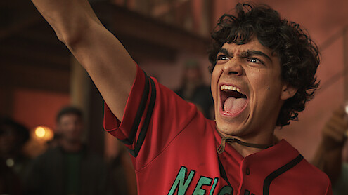 Young man in a red shirt passionately cheering with arm raised in a lively indoor setting, blurred crowd and warm lighting in the background.