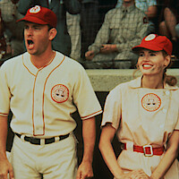 Two baseball players in vintage uniforms stand in a stadium dugout, one shouting with an intense expression and the other smiling, with spectators and netting visible in the background.