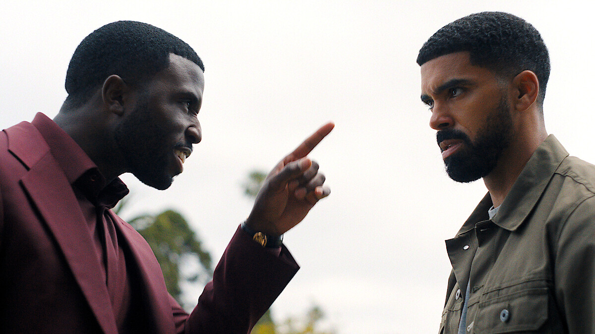 Two men outdoors, one in a maroon suit angrily pointing a finger at another man in a green jacket, who looks serious; trees and cloudy sky in the background.