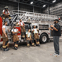 A group of firefighters in uniform listen to a man giving instructions beside a fire truck inside a large, well-lit indoor garage or fire station.