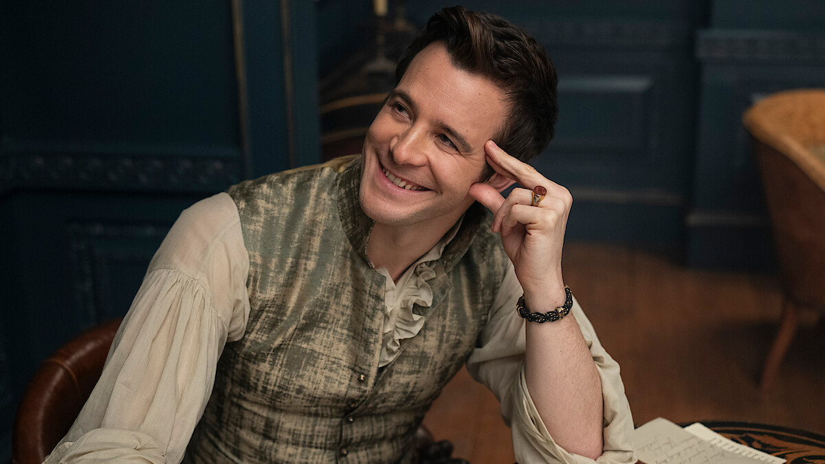 Man in period clothing sitting at a wooden desk, smiling and looking to the side in a warmly lit, elegant room with dark paneling and papers on the desk.