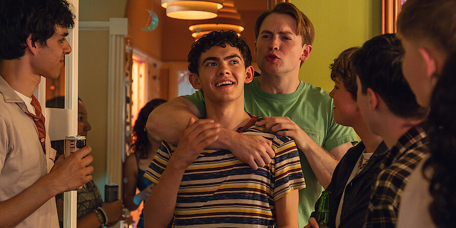 A group of teenagers gather at a lively indoor party. Two boys stand at the center, one smiling while the other playfully wraps an arm around him. The warm, decorated hallway is filled with friends chatting and enjoying themselves.