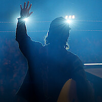 Thomas Rhett stands on stage in silhouette, facing bright spotlights with one hand raised. Smoky blue lighting fills the scene, and the audience is barely visible in the background.