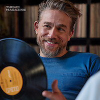 Charlie Hunnam sits in a living room wearing white denim pants and a vintage blue T-shirt spinning a vinyl record in his hands with a smile on his face.