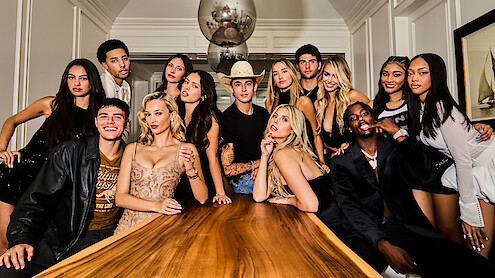 A group of stylish young adults poses around a long wooden table in an elegant, modern room with white walls, artwork, and a decorative light fixture above, capturing a lively and social atmosphere.