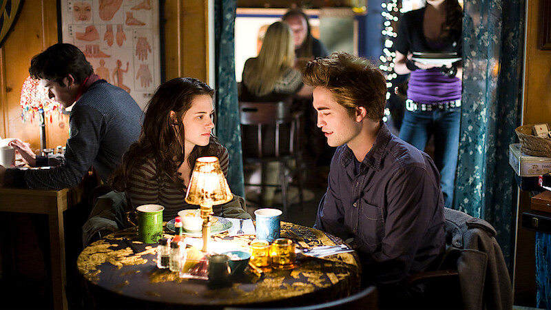 A young woman and man sit at a small round table with drinks and a lamp between them in a cozy, warmly-lit café, talking closely while other people sit and walk in the background.