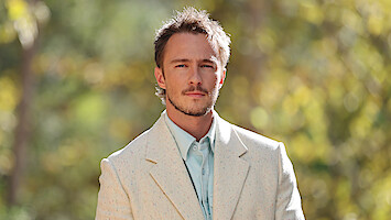 Drew Starkey in a light-colored suit and blue shirt stands outdoors, with soft sunlight and greenery blurred in the background, giving a natural, serene atmosphere.