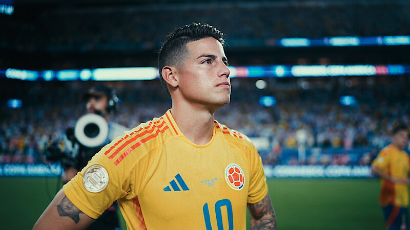A soccer player in a yellow Colombia jersey stands focused on the field, with a stadium full of spectators and media person in the background during a match.