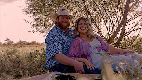 Smiling couple sitting on a blanket outdoors in a grassy field, with trees in the background, wearing casual clothes and cowboy hat, enjoying a relaxed moment in nature during daylight.