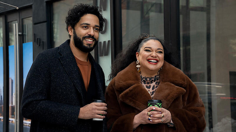 Two people smiling and holding coffee cups, standing outside a modern urban building. The man wears a dark coat and the woman is in a brown fur coat with bold accessories. City reflections are visible in the glass behind them.