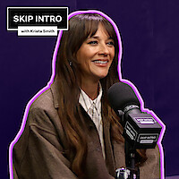 Rashida Jones, sorrindo, sentada diante de um microfone, com um fundo roxo-escuro.