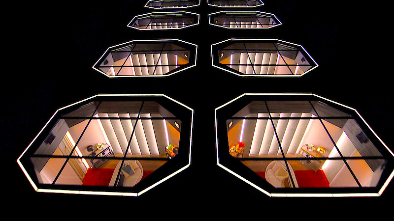 Rows of illuminated, modern geometric windows on a dark building facade at night, each showing stylish interiors with red and orange accents, chairs, and small tables, creating a futuristic architectural pattern.