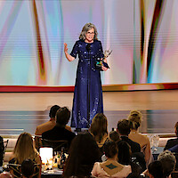 Woman in a blue dress holding an award and speaking on stage at a formal event, with audience members seated at tables in the foreground and colorful abstract background on stage.