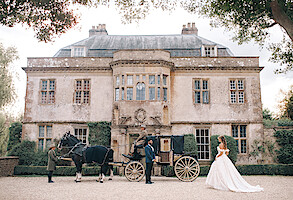Bride in a white gown stands near a vintage horse-drawn carriage outside an elegant, historic mansion. Lush greenery frames the scene, evoking a romantic, fairytale atmosphere. The mood is dreamy and regal.