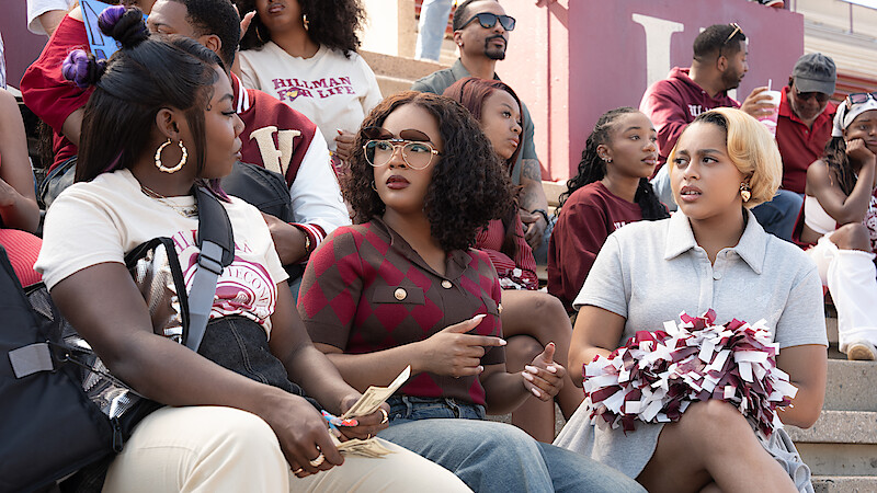 Three women sit on stadium bleachers at a sports event, talking. One holds cash, another has pom-poms. People in the background wear maroon and white, suggesting a school or college environment.