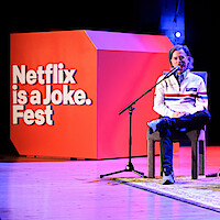 David Spade seated on stage with a microphone, speaking at a Netflix is a Joke Fest event. Bright stage lighting, large red event sign in background, wooden floor and casual atmosphere.