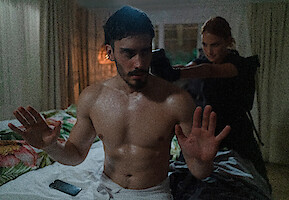 A man in a towel holds up his hands as a woman in uniform points a gun at the back of his head.