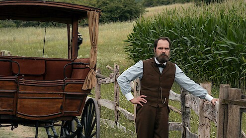 A man in period clothing stands by a wooden fence next to an old carriage in a rural setting with green cornfields and meadows in the background.