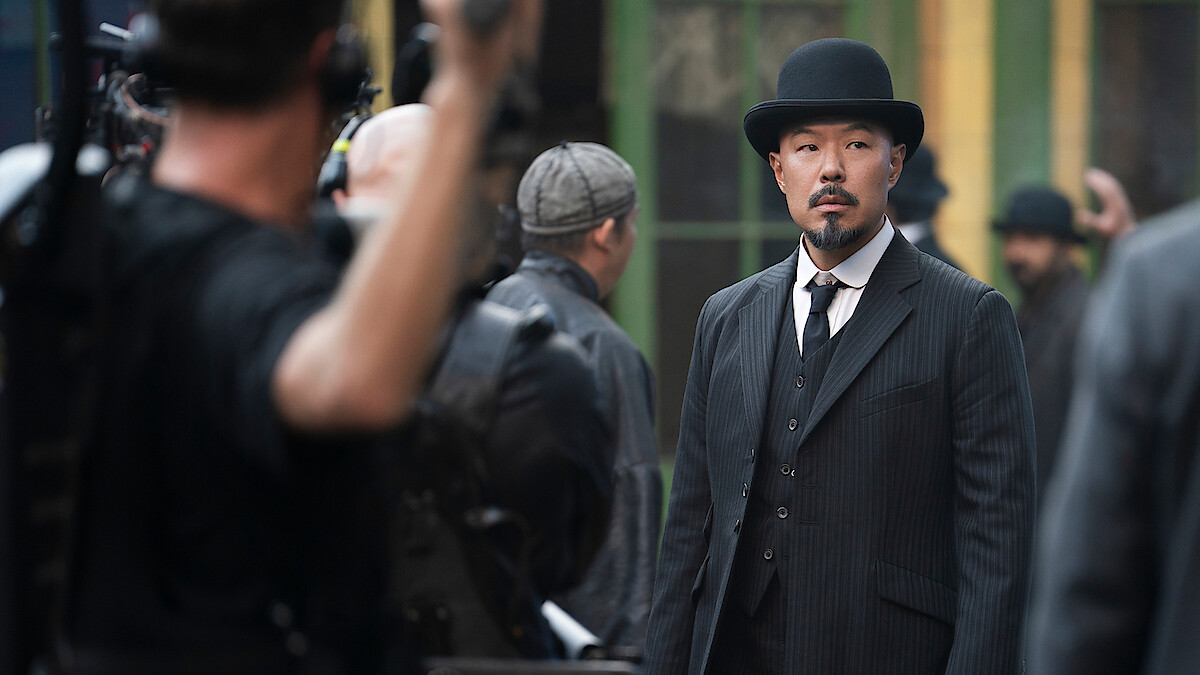 Man in a black suit and bowler hat stands outside on a film set, surrounded by people in period clothing, with cameras and crew visible, suggesting a historical or dramatic movie production being filmed.