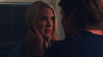 A blonde woman looks anxious as a man gently holds her face in a dimly lit room, suggesting a tense or emotional moment between the two people indoors at night.