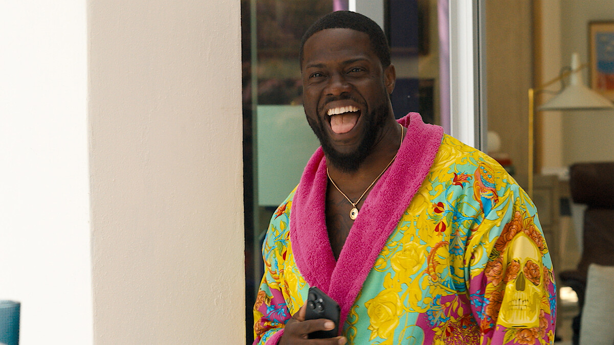 Man standing indoors near a doorway, laughing joyfully, wearing a colorful bathrobe with a bright pink collar and holding a smartphone, with modern home furnishings in the background.