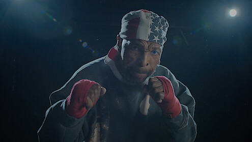 A male boxer wearing red hand wraps and a USA flag bandana poses in a fighting stance under dramatic lighting in a dark gym or training environment.