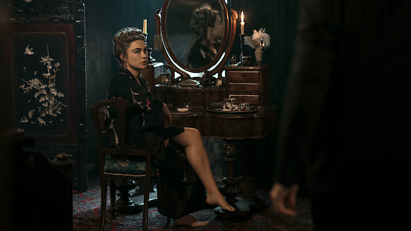 Woman in a dark robe sits at a vintage vanity with candles and flowers in a dim, elegant room, looking toward a standing figure partially visible in the foreground.