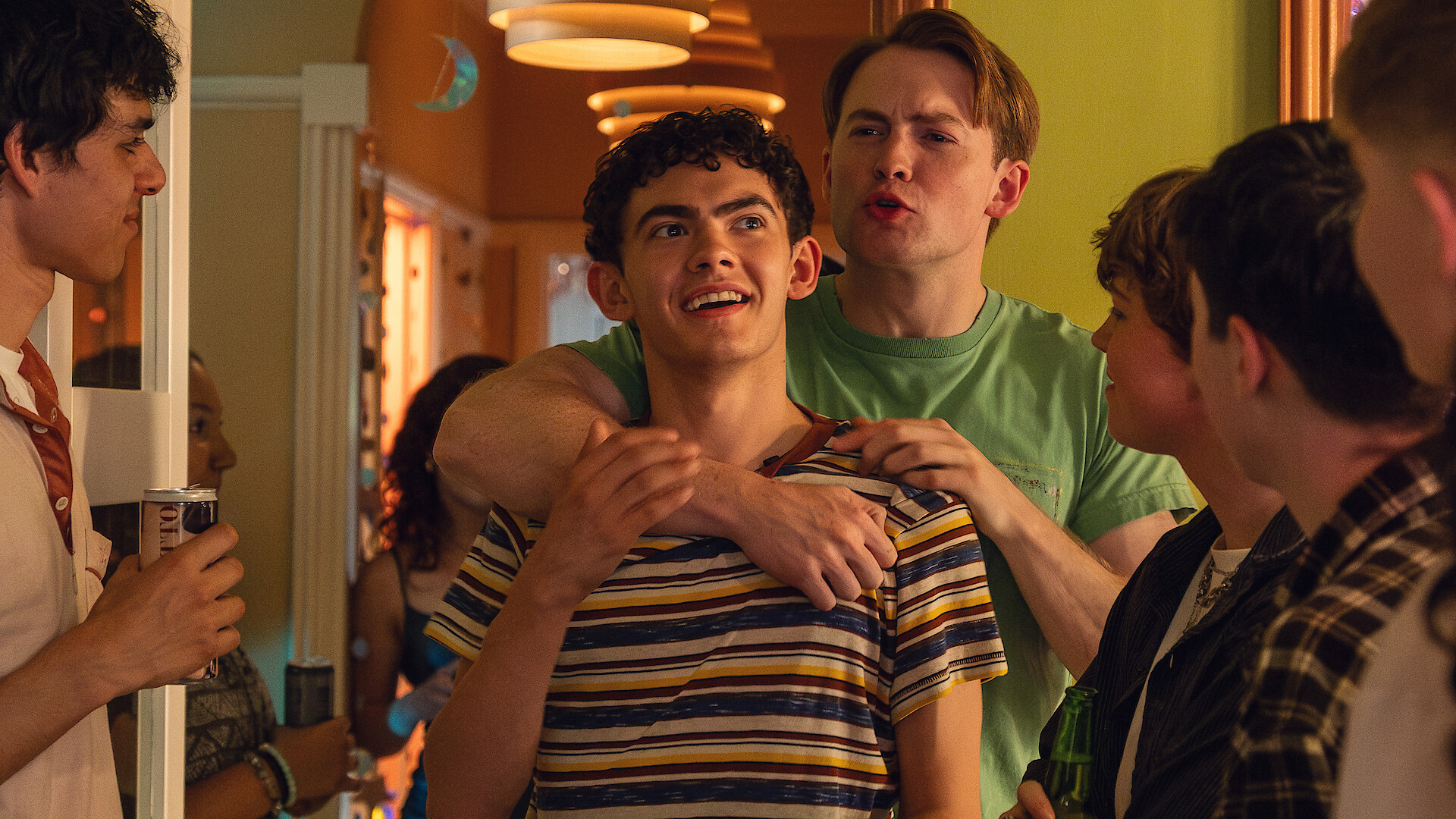 A group of young people at an indoor party, with one person playfully embracing another from behind while others watch and socialize under warm lighting.