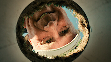 A smiling man wearing a white headband is looking down into a hole, possibly on a golf course, with sunlight and blue sky in the background, viewed from inside the hole.