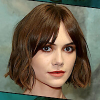 A person with short brown hair and bangs, neutral expression, photographed in a studio setting with a muted, artistic background.