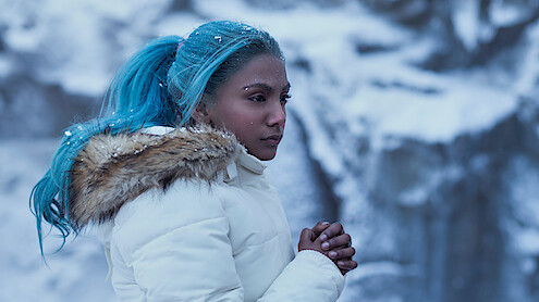 Charithra Chandran as Princess Vivi, with blue hair in a white winter coat stands outdoors in a snowy, icy landscape, looking thoughtful and cold with hands clasped, snowflakes in hair and on coat.