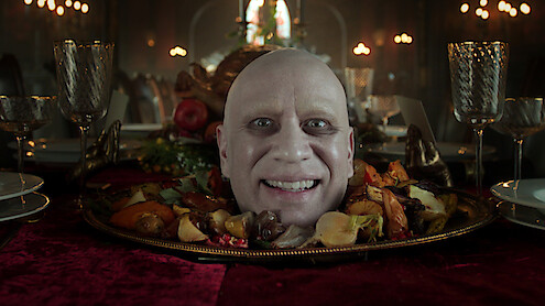 A smiling bald man’s head appears on a platter surrounded by roasted vegetables, set on an elegant dining table with ornate glasses and a dimly lit, candlelit gothic banquet hall in the background.