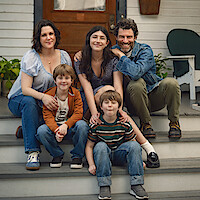 A family of five, including two adults, a teen girl, and two young boys, sits together smiling on the front porch steps of a house with white siding, plants, and a wooden door, suggesting a warm, casual outdoor setting.