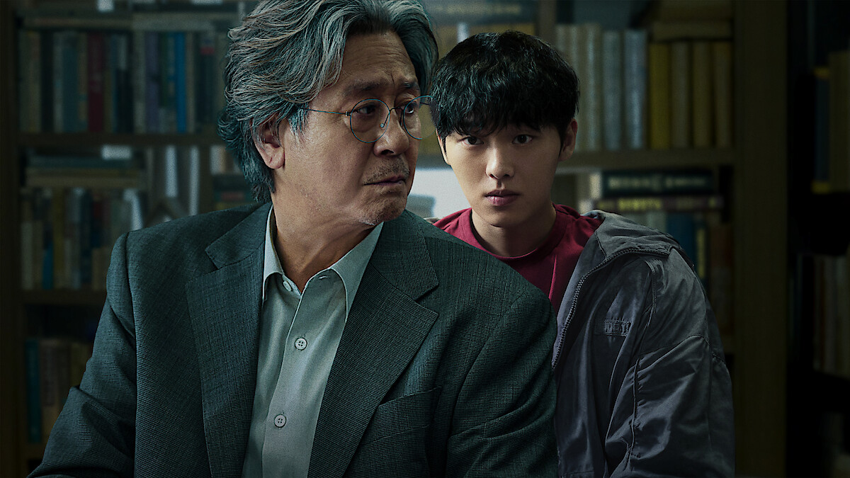 Two men, one older with gray hair in a suit and one younger with dark hair in casual clothes, are in a library or study filled with books, both appearing serious and focused.