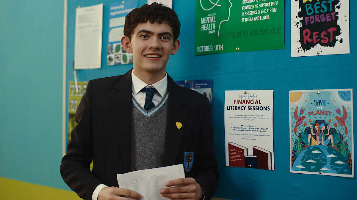 A student in a school uniform stands indoors holding papers, smiling in front of a blue bulletin board with posters about mental health, financial literacy, and environmental awareness.