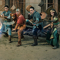 Four young warriors in fantasy costumes face an opponent producing blue energy in an ancient, dimly lit building with oriental decor and scattered debris, suggesting a tense magical battle scene.
