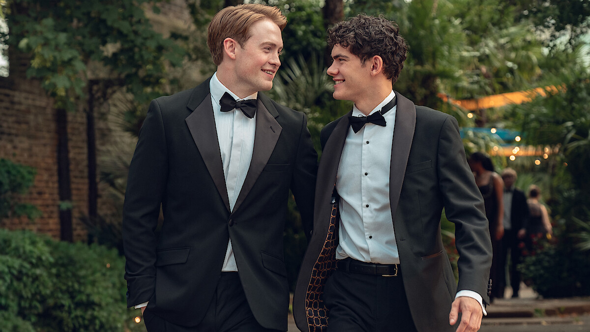 Two young men in tuxedos walk outdoors at an evening event, smiling at each other, surrounded by greenery and string lights in a festive setting.