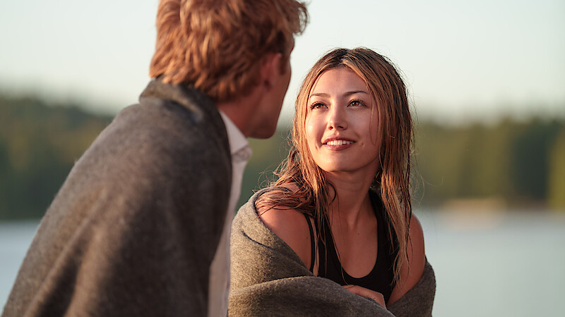 Two people wrapped in a blanket sit by a calm lake at sunset, conversing and smiling, with trees in the background and soft natural lighting.