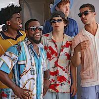 Five men wearing colorful shirts and sunglasses stand together outdoors, laughing and talking in a casual, sunny setting.