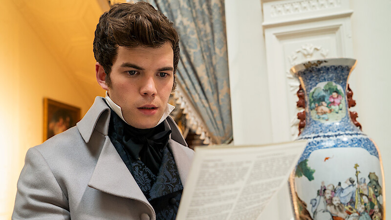 A young man in period costume reads a letter or paper indoors, standing near a large ornate vase with a decorative pattern in an elegant room with classic furnishings and drapery.