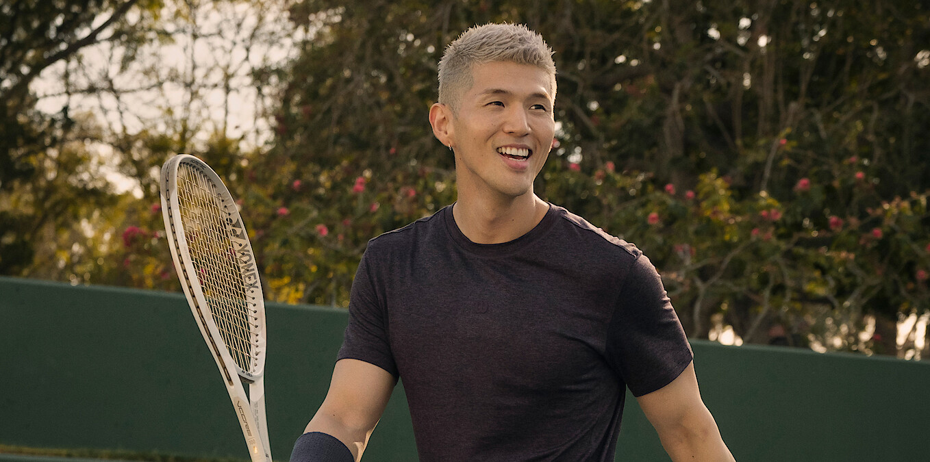 Smiling person with short light hair holding a tennis racquet on an outdoor tennis court, wearing a dark shirt, with trees and greenery in the background.