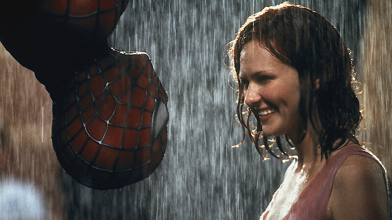 Spider-Man hangs upside down in the rain, about to kiss a smiling woman who stands close to him, both illuminated and wet, in an urban night setting.