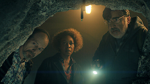 Three people look down into a hole inside a dimly lit cave or tunnel, one holding a flashlight, with an exposed bulb overhead and rocky surroundings, creating a mysterious and tense atmosphere.