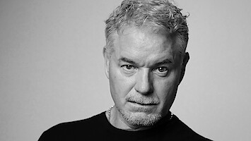Eric Dane de cabelos curtos, claros e cacheados e barba, vestindo uma camisa preta, em pé diante de um fundo liso de cor clara e com expressão séria; foto em preto e branco com iluminação de estúdio.