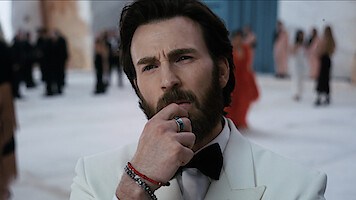 A thoughtful man in a white tuxedo touches his chin at a formal indoor event, with blurred people in elegant attire in the background.